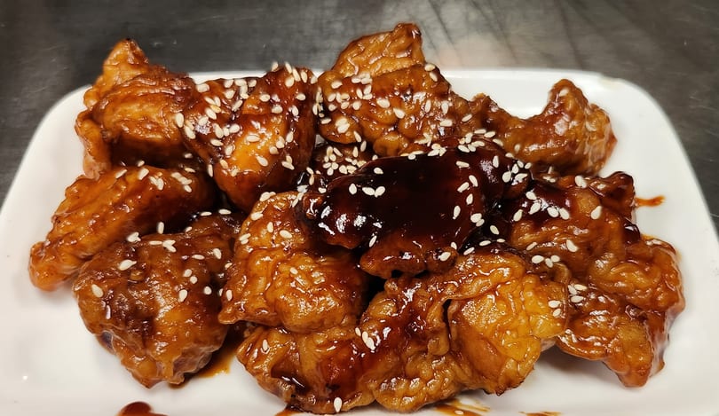 Close-up of sesame chicken pieces coated in a glossy brown sauce on a white plate.